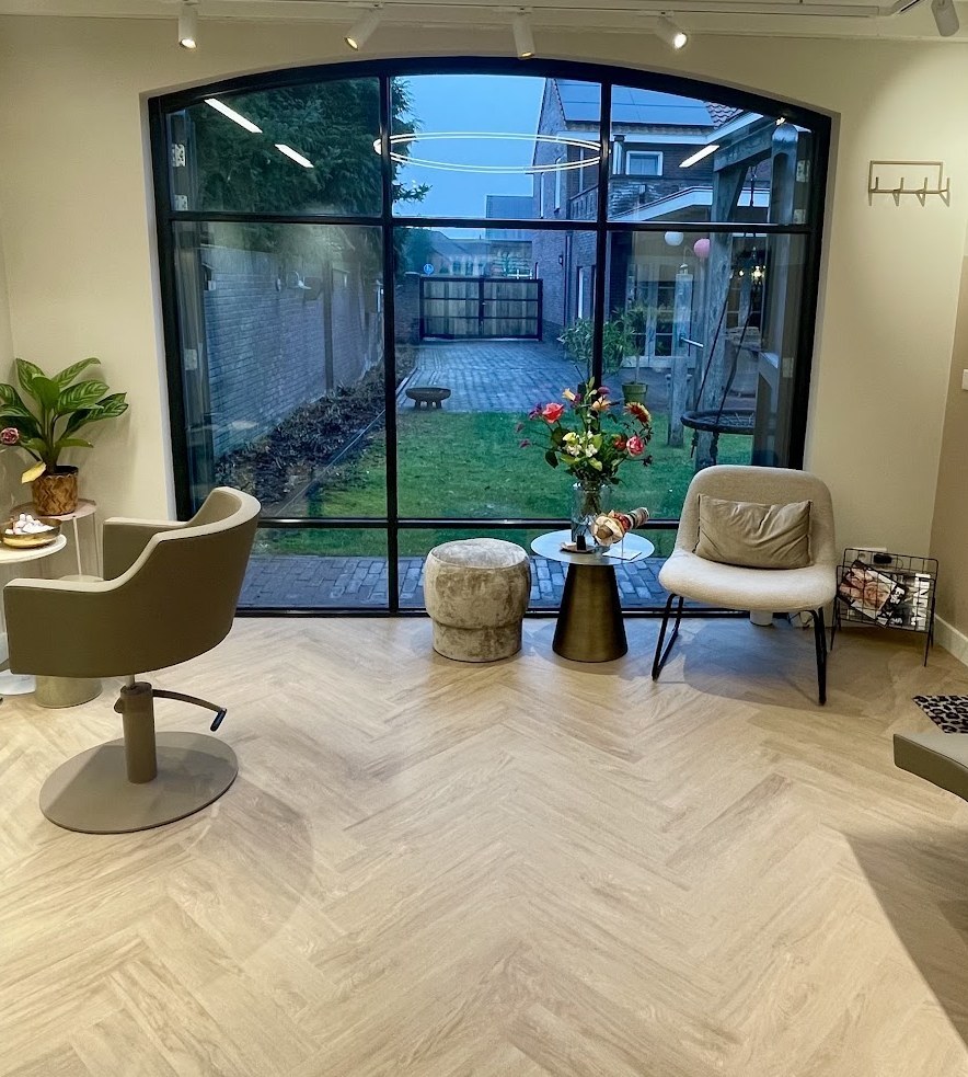 Groot stalen raam met boog en uitzicht op de tuin, met kappersstoelen en een fauteuil in de salon